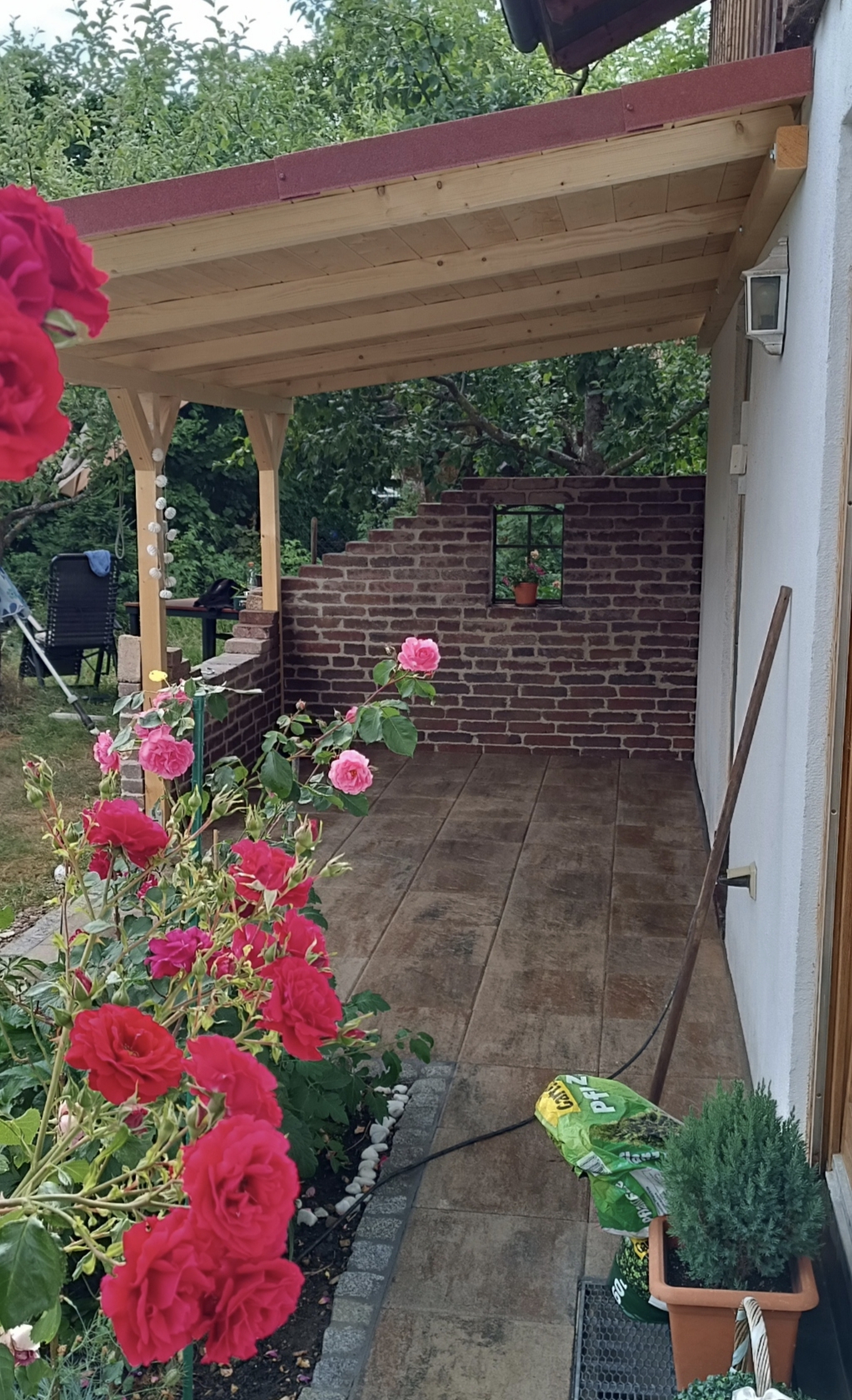 Überdachte Terrasse mit Natursteinfliesen, Backsteinmauer und Rosenbeet